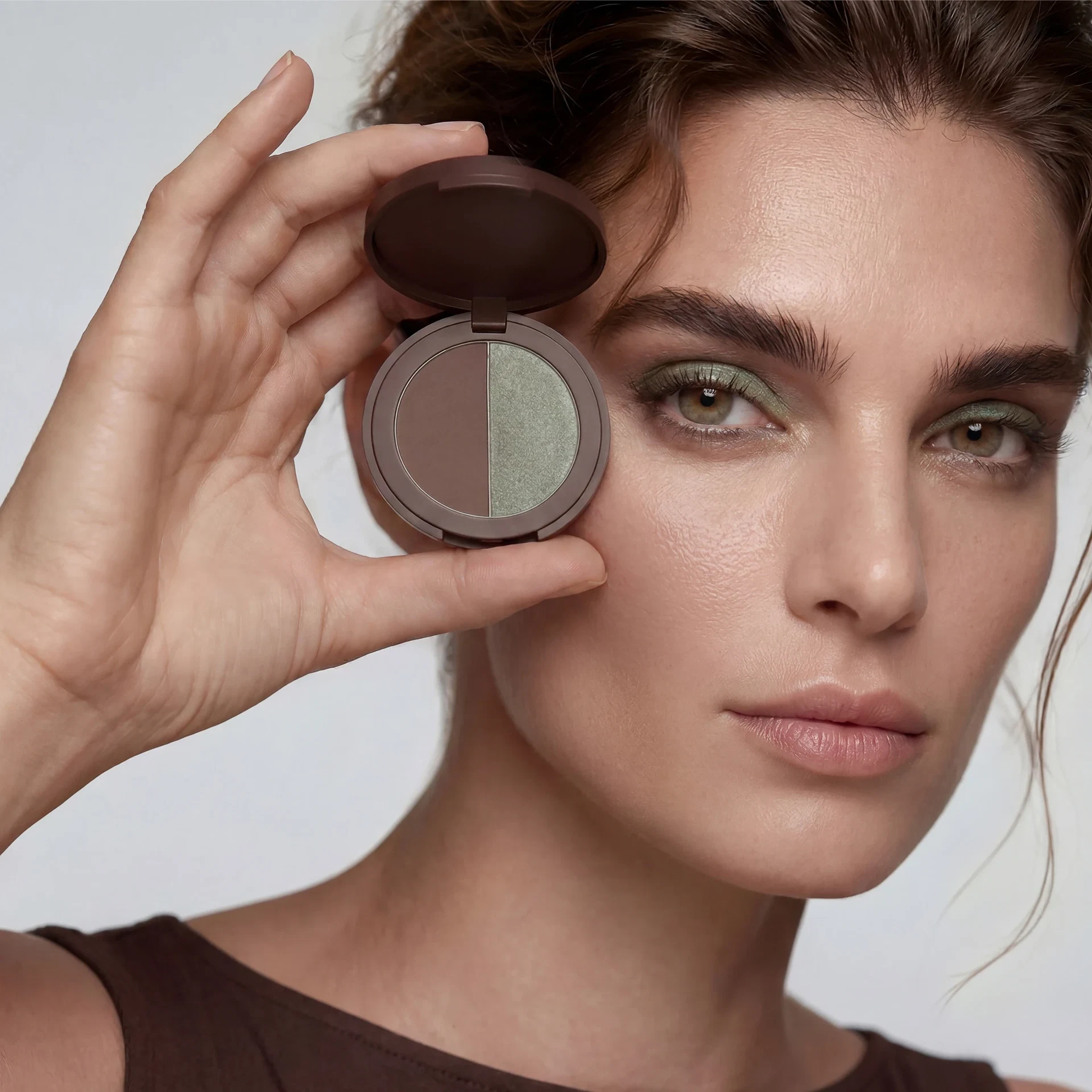Frau mit moosgrünem Augen-Make-up hält eine geöffnete runde Lidschattenpalette mit Moosgrün und Braun neben ihr Gesicht.