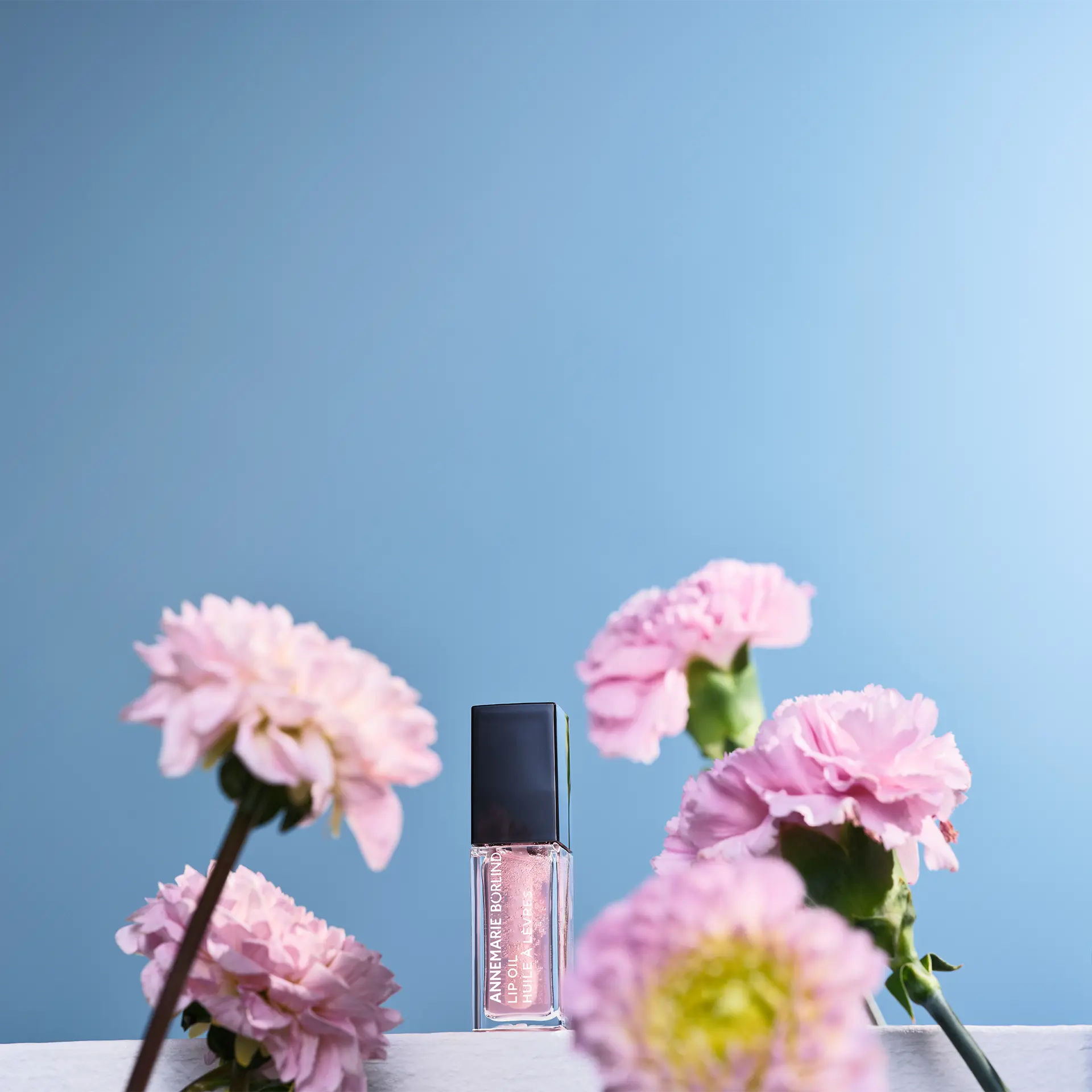 ANNEMARIE BÖRLIND Lip Oil Cherry Blossom in dunklem Beerenton vor blauem Hintergrund zwischen Blumen