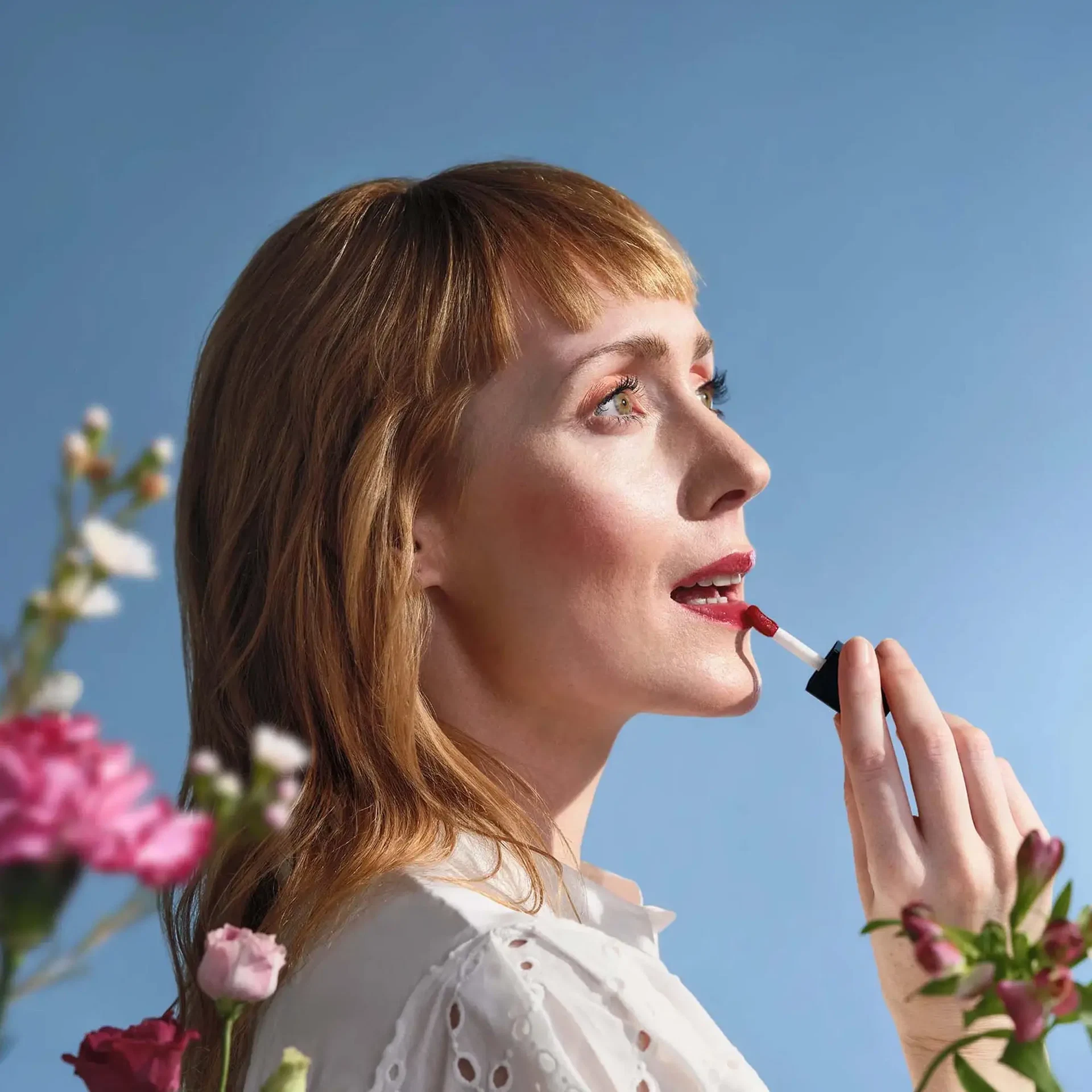Frau trägt das LIP OIL Midnight Dahlia von ANNEMARIE BÖRLIND auf, umgeben von Blüten vor blauem Himmel