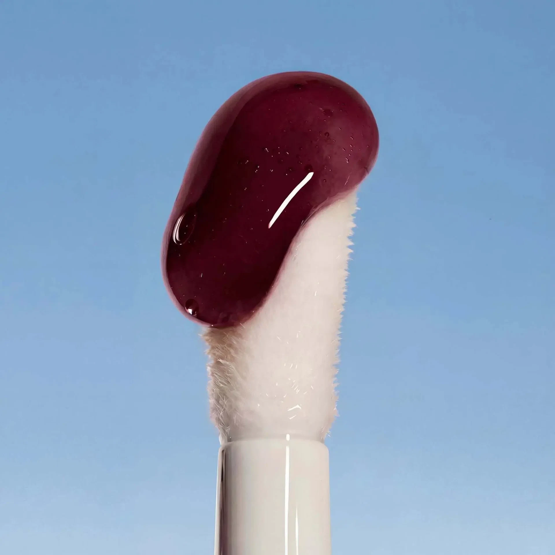 Applikator des LIP OIL Midnight Dahlia mit glänzender, tiefroter Textur vor blauem Hintergrund