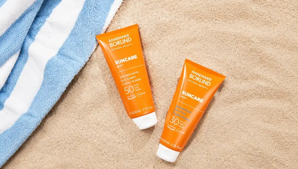 Zwei ANNEMARIE BÖRLIND Sonnencremes mit LSF 50 und LSF 30 im Sand mit gestreiftem Badehandtuch