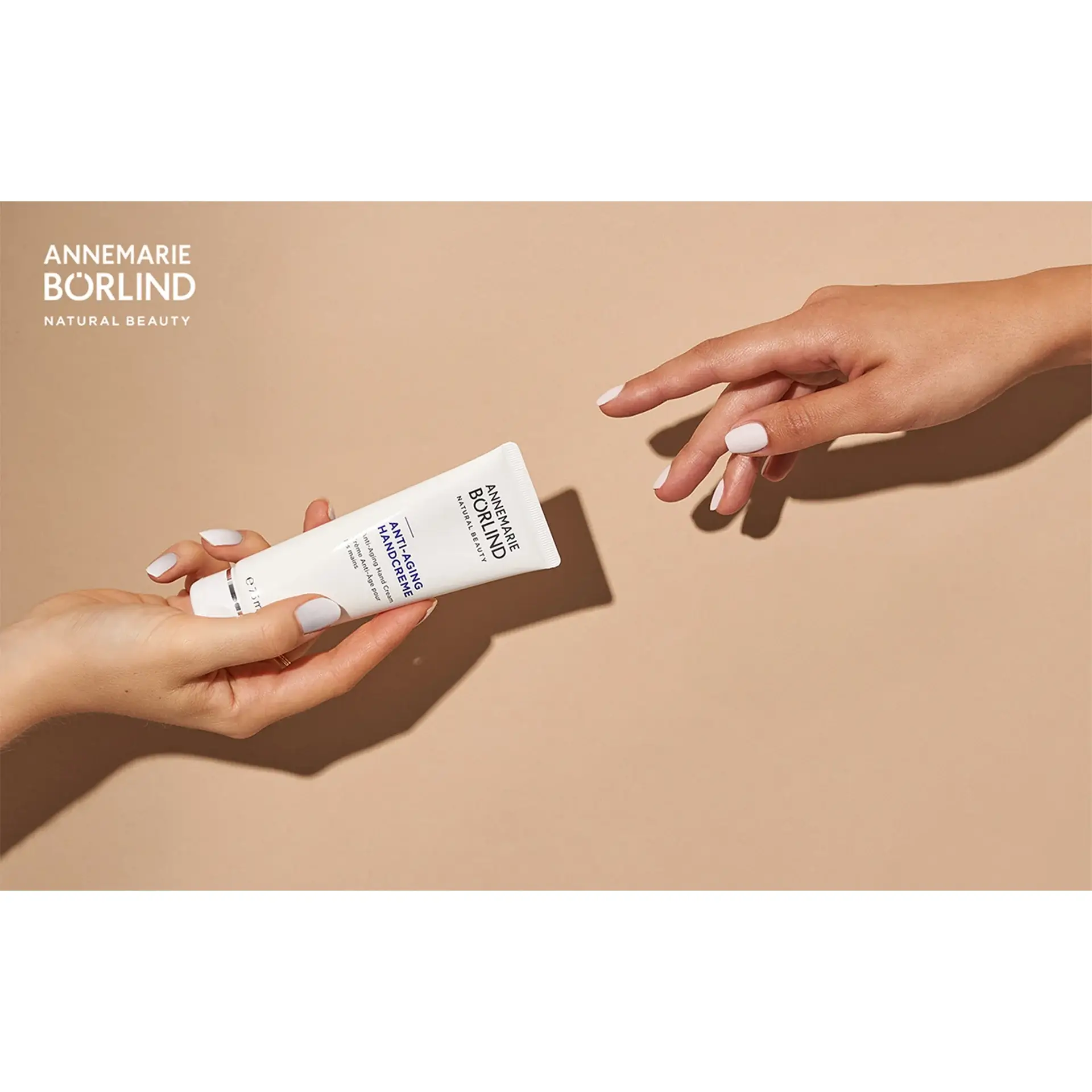 ANNEMARIE BÖRLIND Anti-Aging Handcreme, eine Hand reicht die Tube an eine andere Hand mit beigem Hintergrund