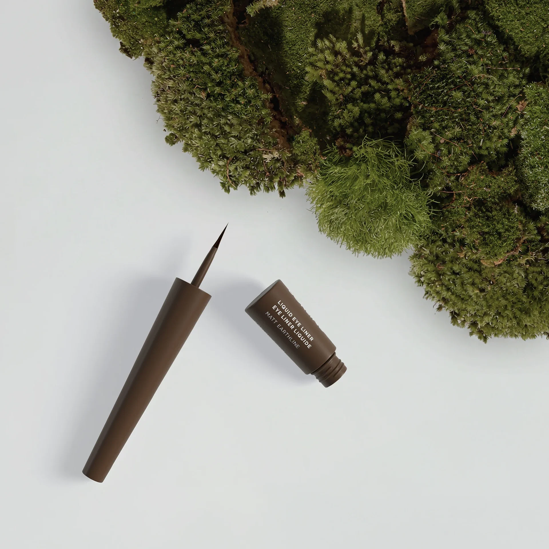 Geöffneter brauner Liquid Eyeliner mit feiner Pinselspitze, arrangiert neben üppigem grünem Moos auf hellem Hintergrund.