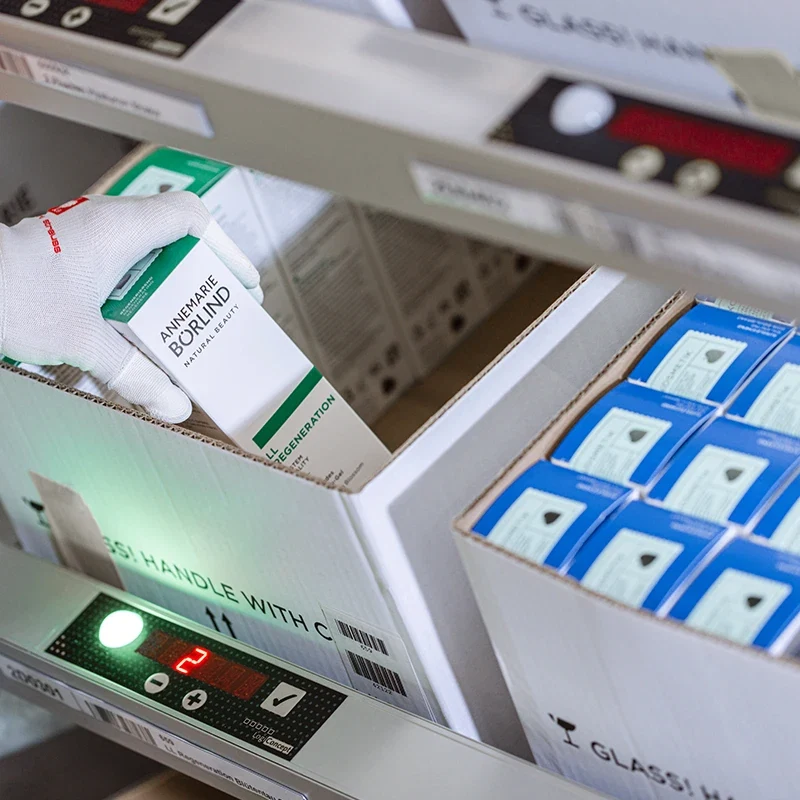 Hand mit Handschuh greift ANNEMARIE BÖRLIND Produktverpackung aus Versandkarton an moderner Kommissionieranlage