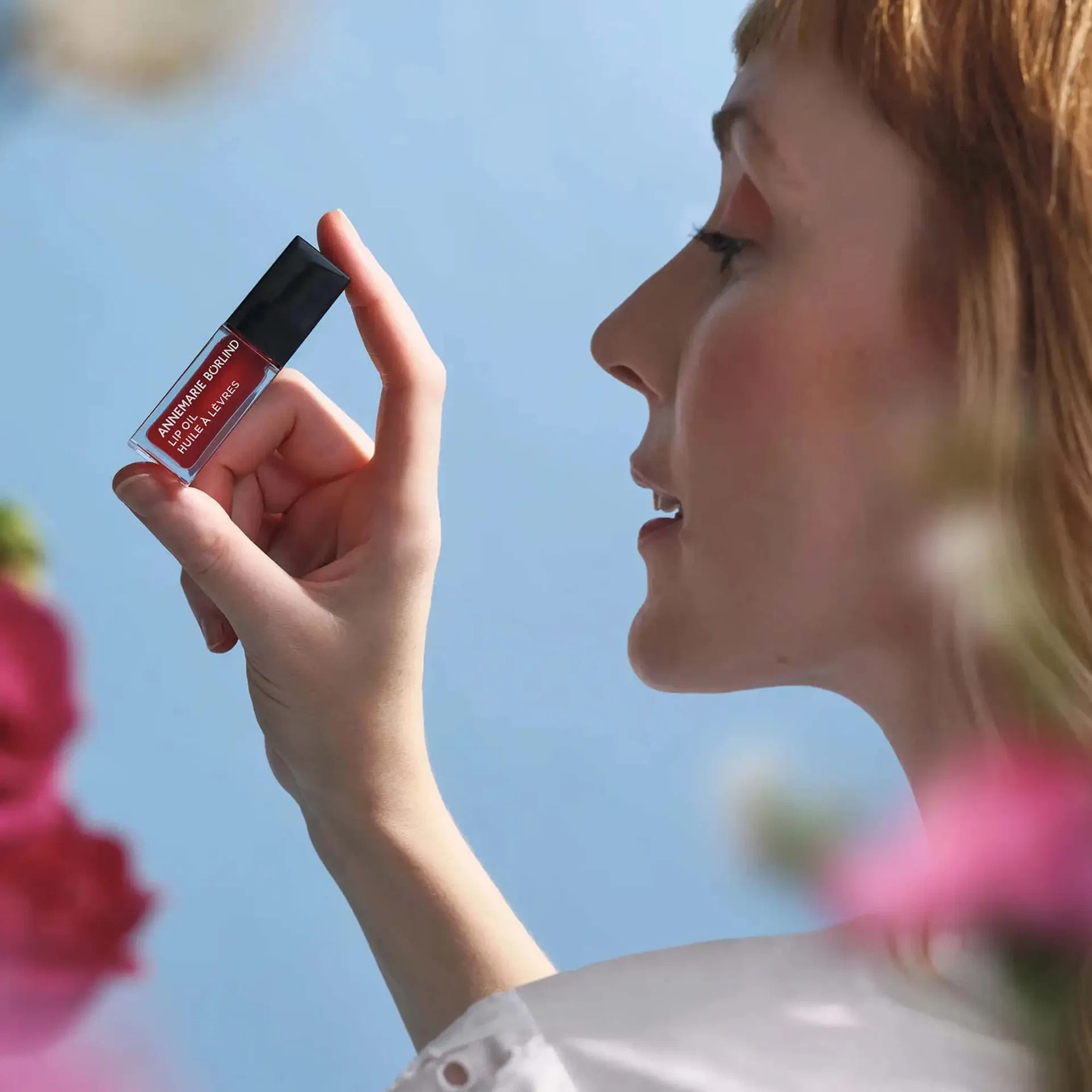Frau hält das LIP OIL Wild Fuchsia von ANNEMARIE BÖRLIND vor blauem Himmel, umgeben von Blüten
