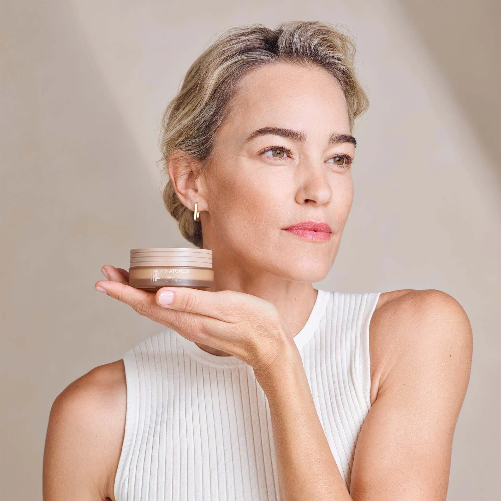 Frau hält NATUNEO Refining Day Cream in Hand, blickt nach rechts, trägt weißes ärmelloses Oberteil