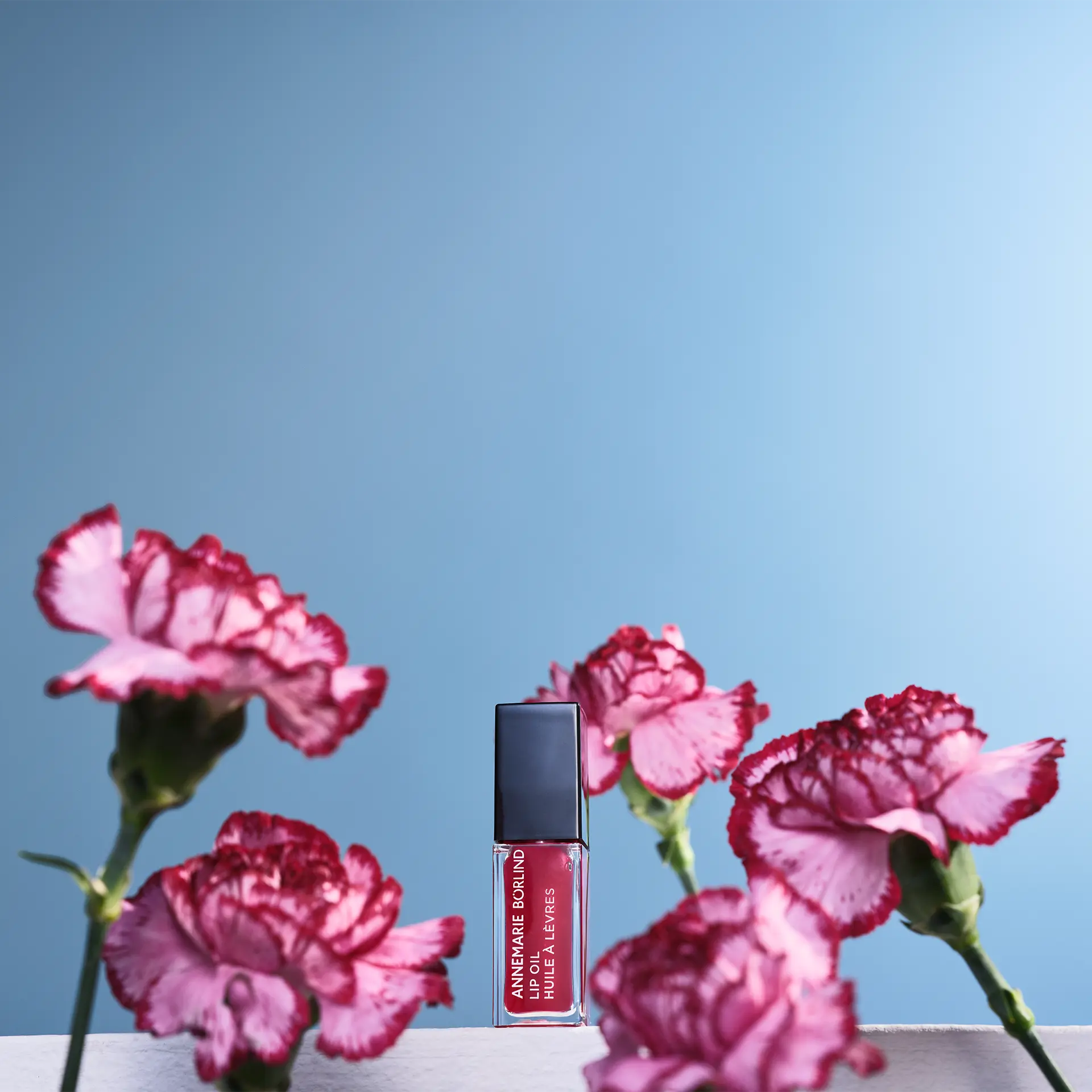 ANNEMARIE BÖRLIND Lip Oil Wild Fuchsia in mittlerem Beerenton vor blauem Hintergrund zwischen Blumen