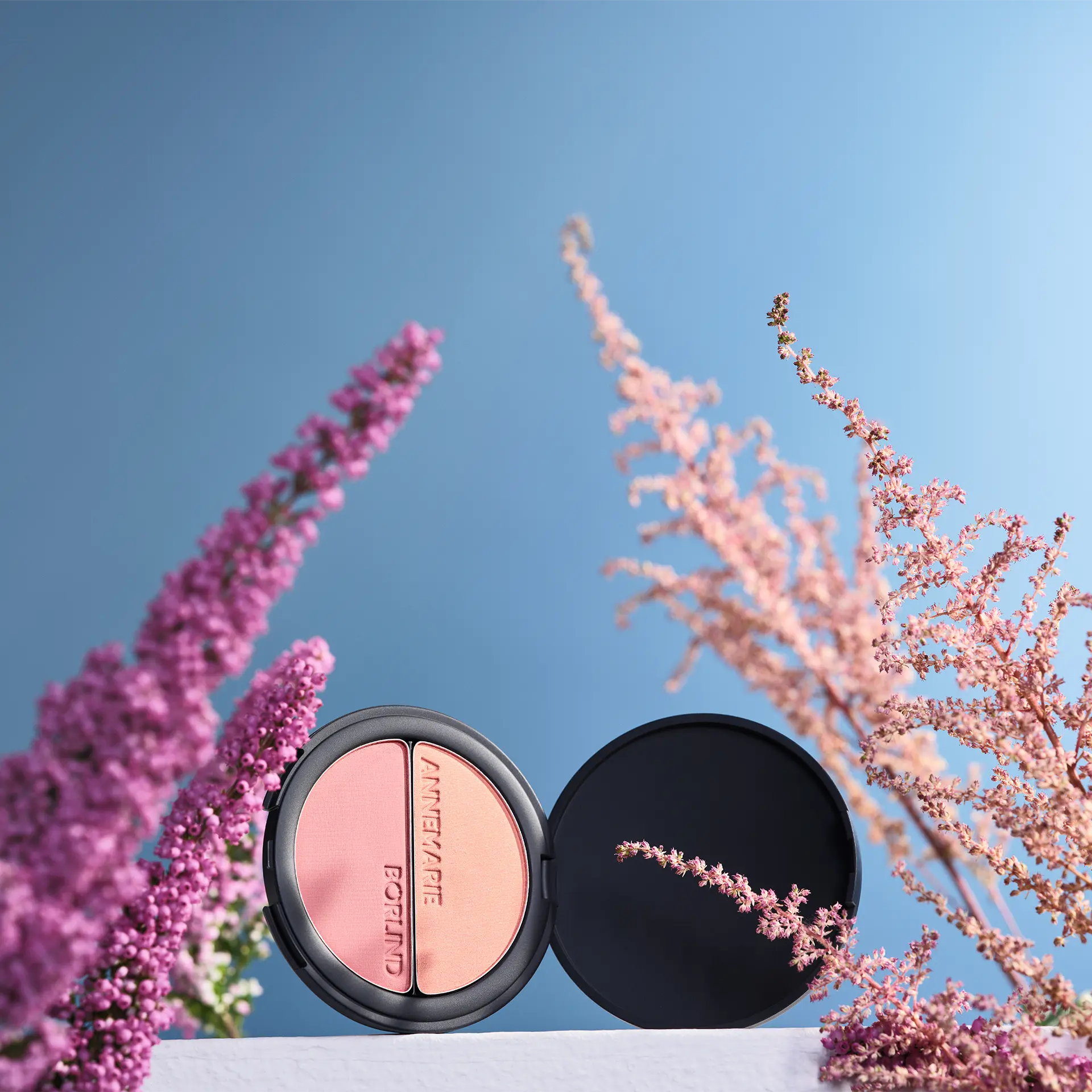 ANNEMARIE BÖRLIND Duo Blush & Eye Shadow in schwarzer Dose mit Logo vor blauem Hintergrund zwischen Blumen