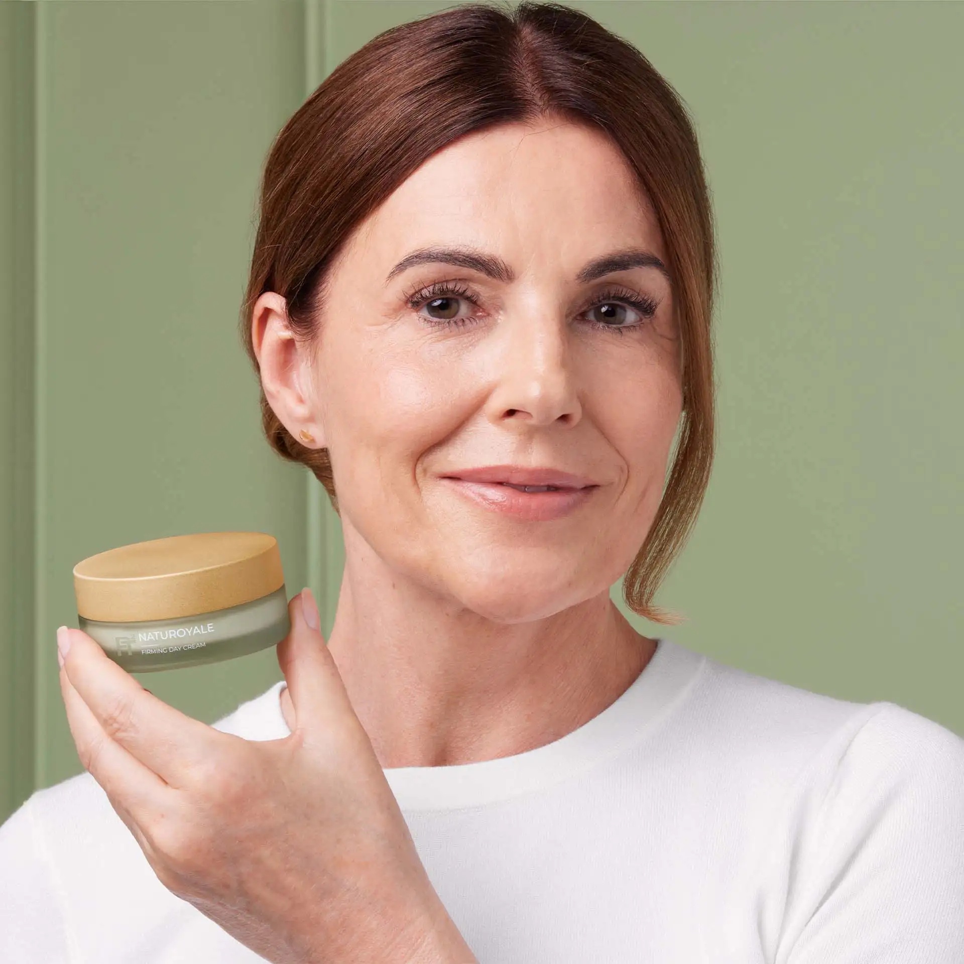 Frau hält NATUROYALE Firming Day Cream, lächelt leicht, grüner Hintergrund, weiße Bluse