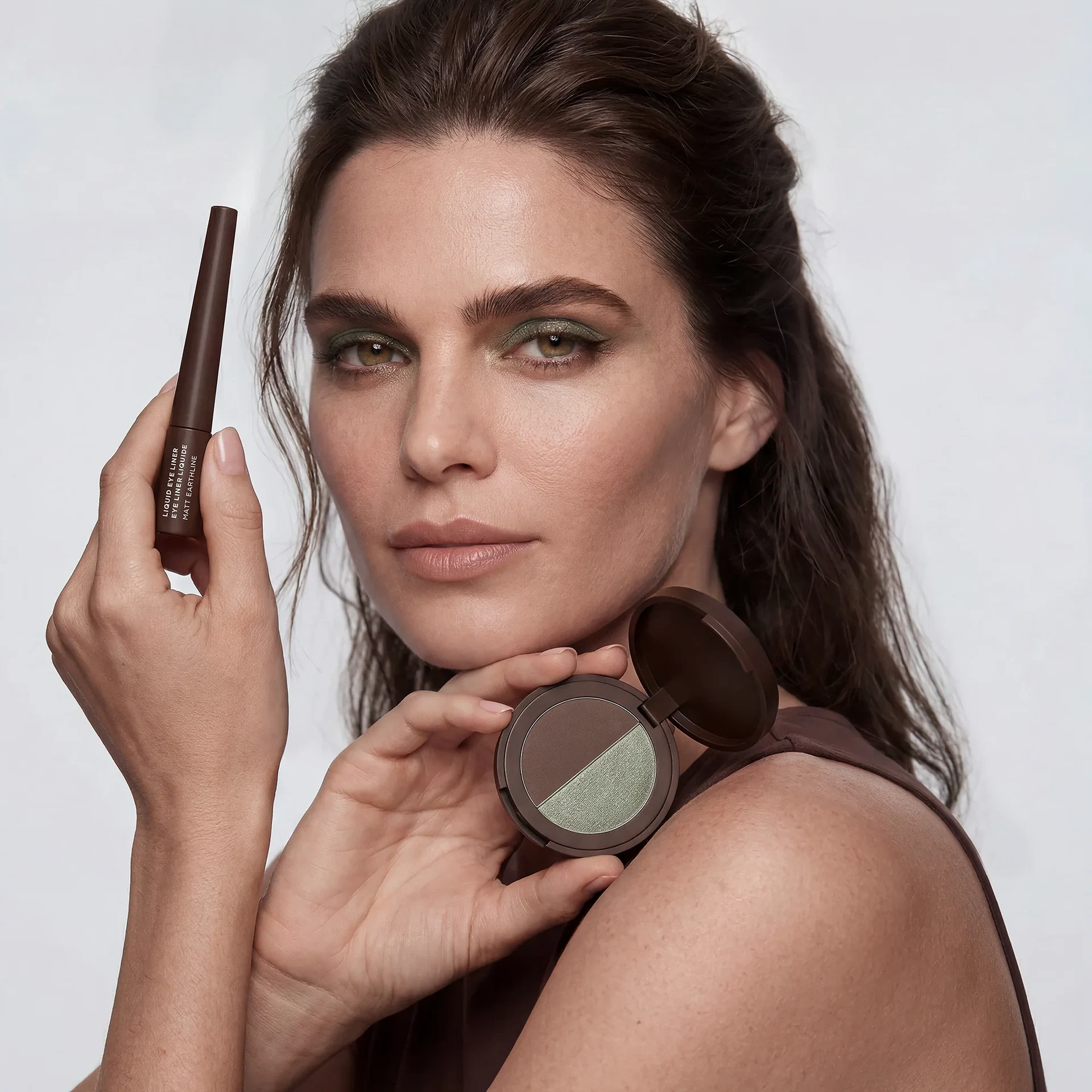 Frau mit moosgrünem Augen-Make-up hält einen braunen Liquid Eyeliner und eine geöffnete Lidschattenpalette in Braun und Moosgrün.