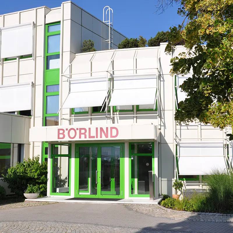 Eingang des Firmensitzes von BÖRLIND mit grünen Türen, Sonnenschutz und Begrünung an einem sonnigen Tag