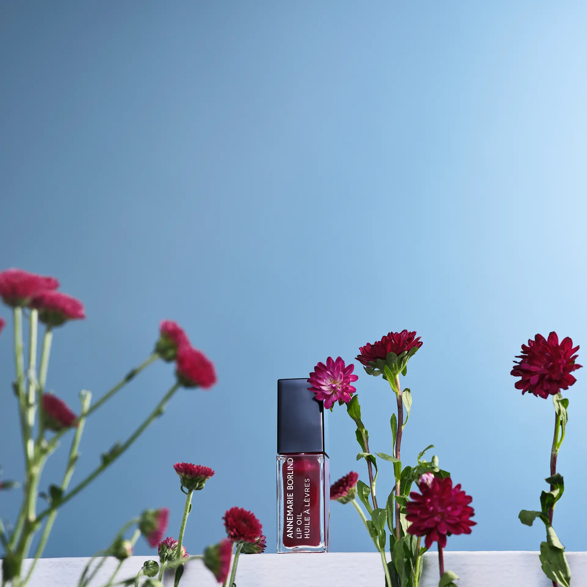 ANNEMARIE BÖRLIND Lip Oil Midnight Dalia in dunklem Beerenton vor blauem Hintergrund zwischen Blumen