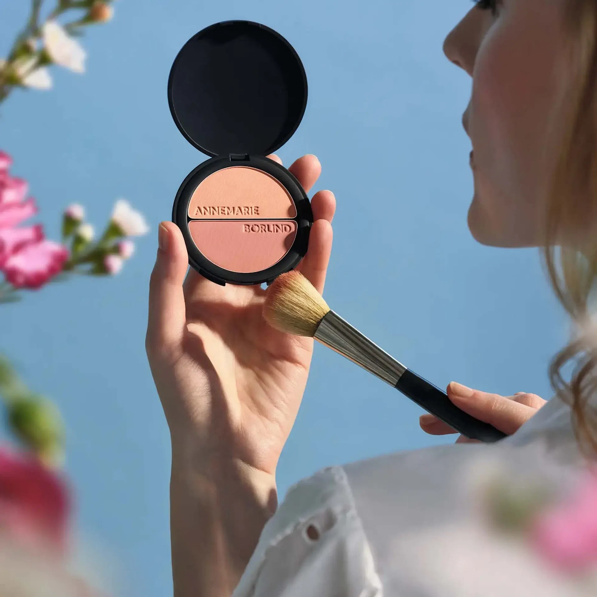 Frau hält das geöffnete DUO BLUSH & EYE SHADOW von ANNEMARIE BÖRLIND in Pfirsich und Rosé und nimmt Produkt mit einem Pinsel auf, vor blauem Himmel und Blüten