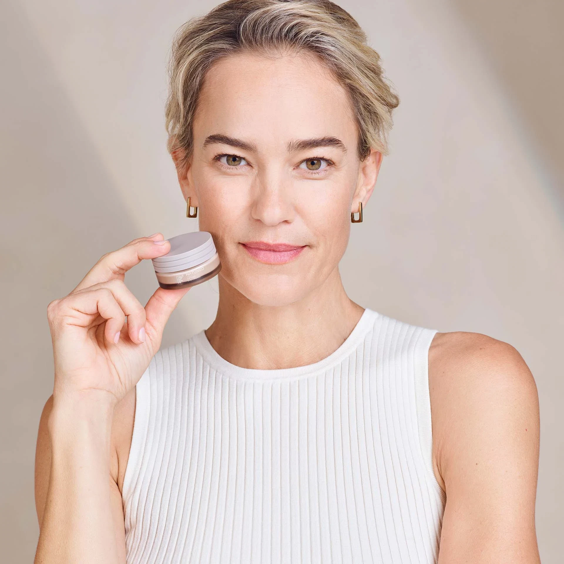 Frau hält NATUNEO Lifting Eye Cream in Hand, trägt weißes Top, neutraler Hintergrund, Blick zur Kamera