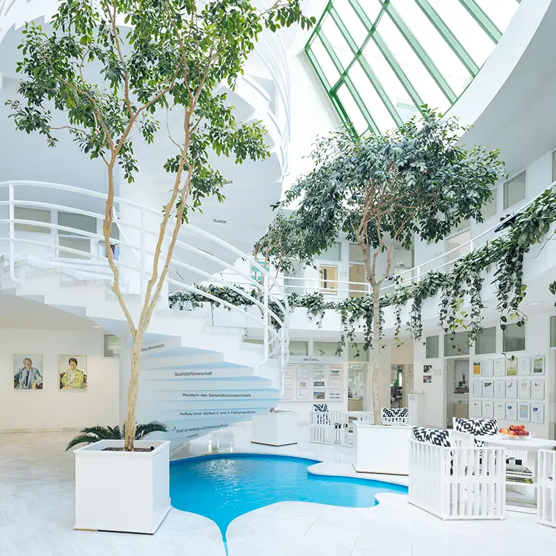 Lichtdurchflutetes Atrium des BÖRLIND-Firmensitzes mit begrünten Innenbäumen, Wasserbecken und weißer Wendeltreppe