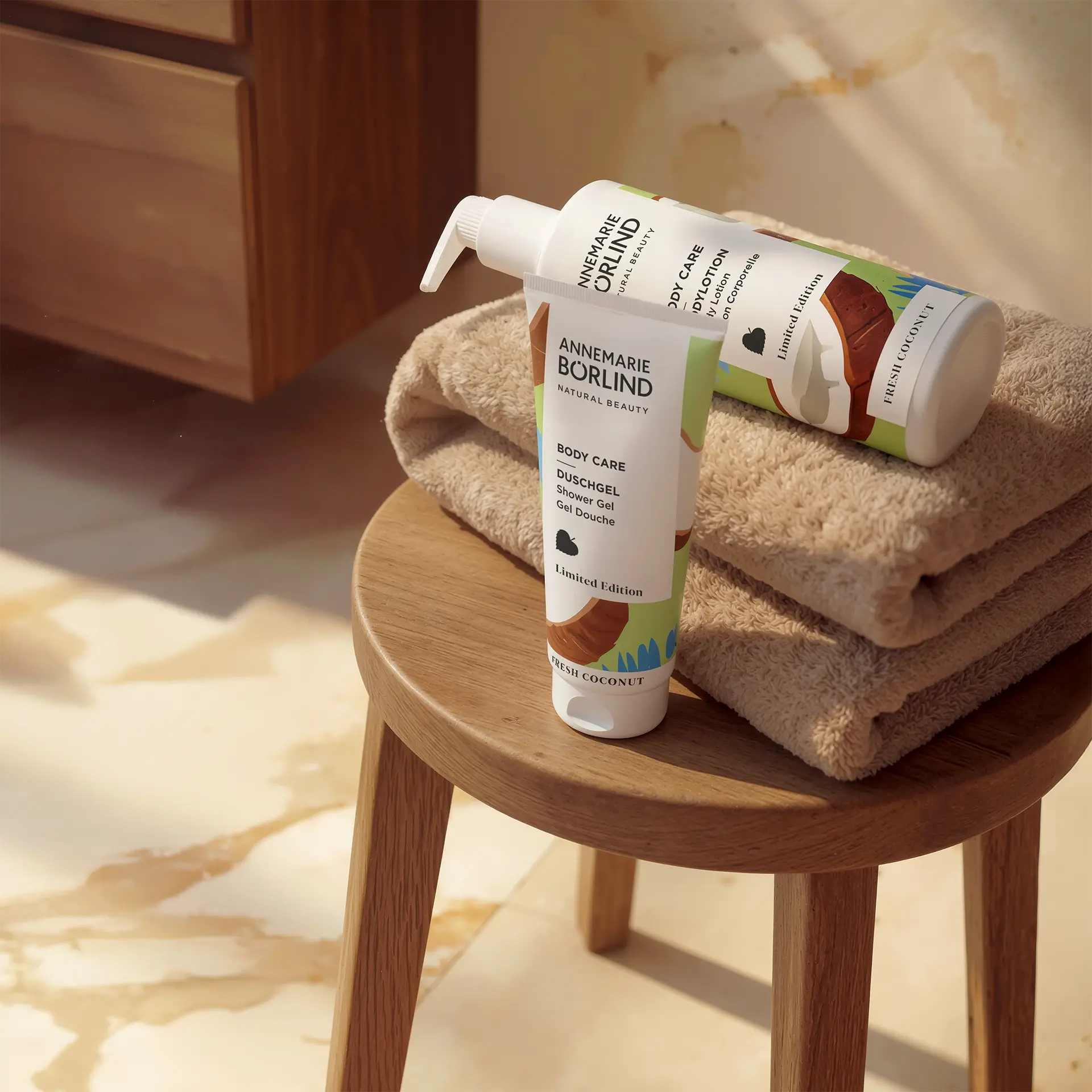 BODY CARE Duschgel und Bodylotion Fresh Coconut mit sommerlichem Design auf braunen Handtüchern und Holzstuhl im Bad