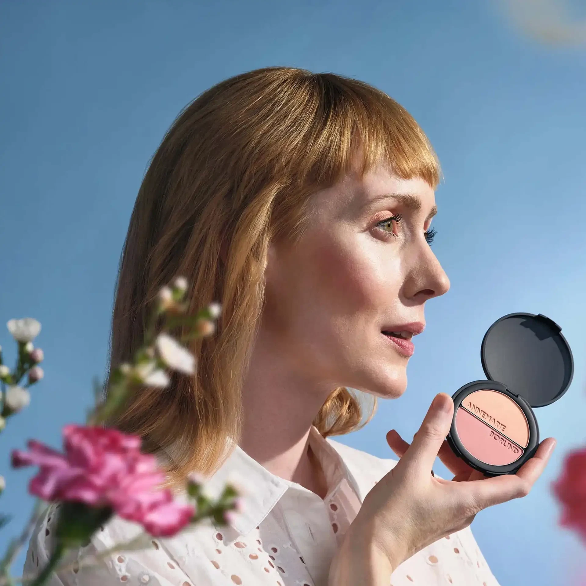 Frau mit frischem Frühlings-Make-up hält das geöffnete DUO BLUSH & EYE SHADOW von ANNEMARIE BÖRLIND in Rosé und Pfirsich vor blauem Himmel, umgeben von Blüten