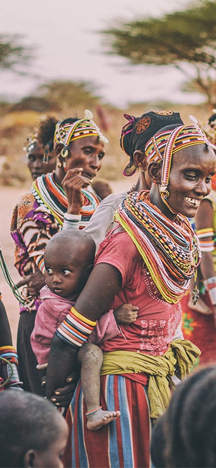 Frauen mit traditioneller bunter Kleidung und Schmuck, eine trägt ein Kind auf dem Rücken, im Freien