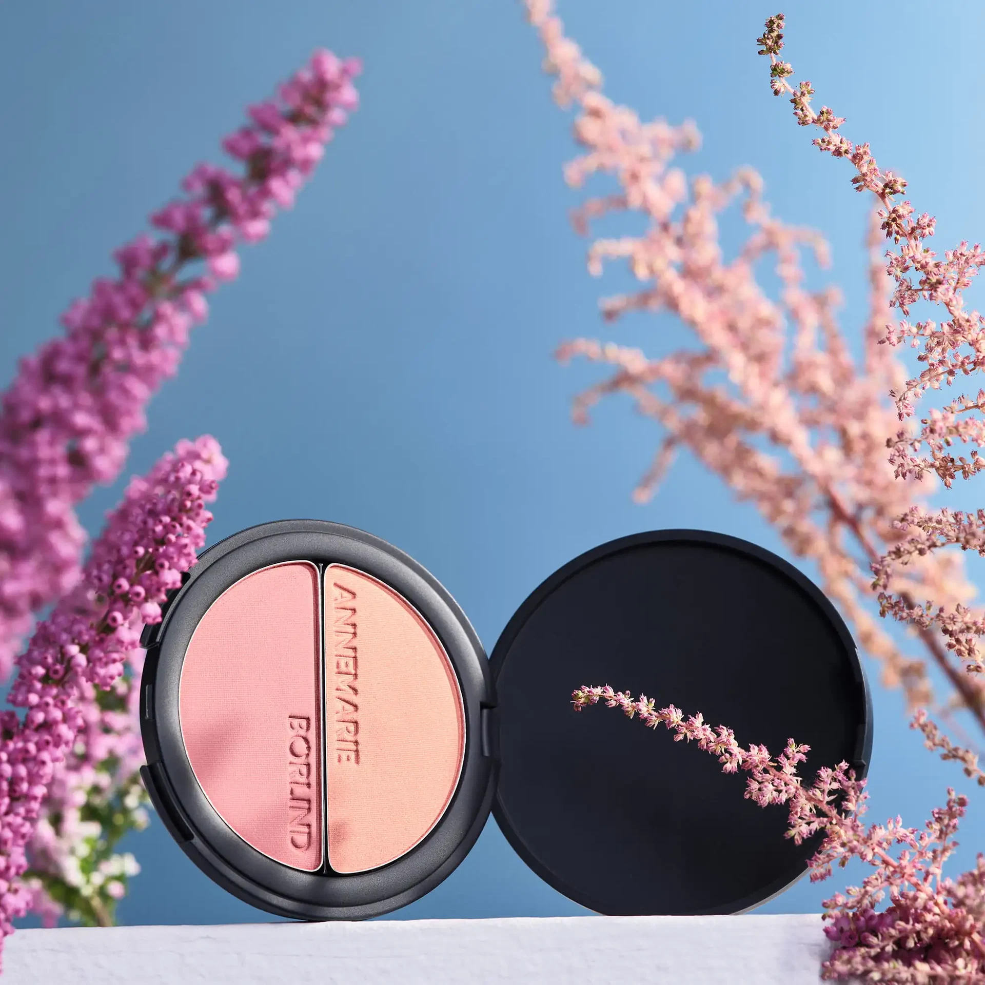 Geöffnetes DUO BLUSH & EYE SHADOW von ANNEMARIE BÖRLIND in Rosé und Pfirsich vor blauem Himmel, umgeben von pinken Blütenzweigen