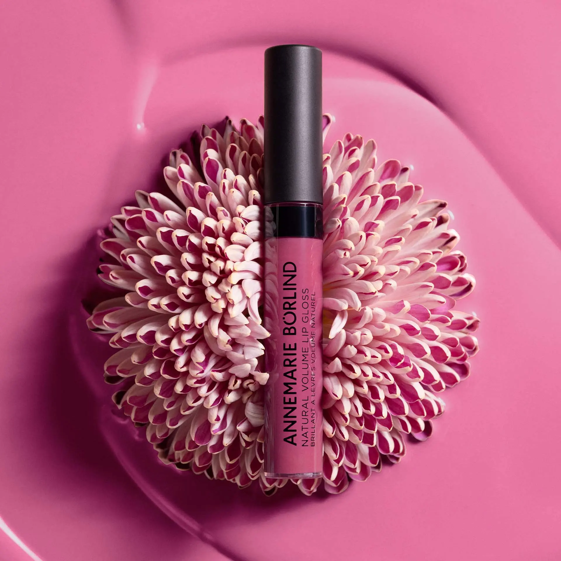 ANNEMARIE BÖRLIND Natural Volume Lip Gloss in Pink mit schwarzem Deckel auf Blume mit Textur als Hintergrund