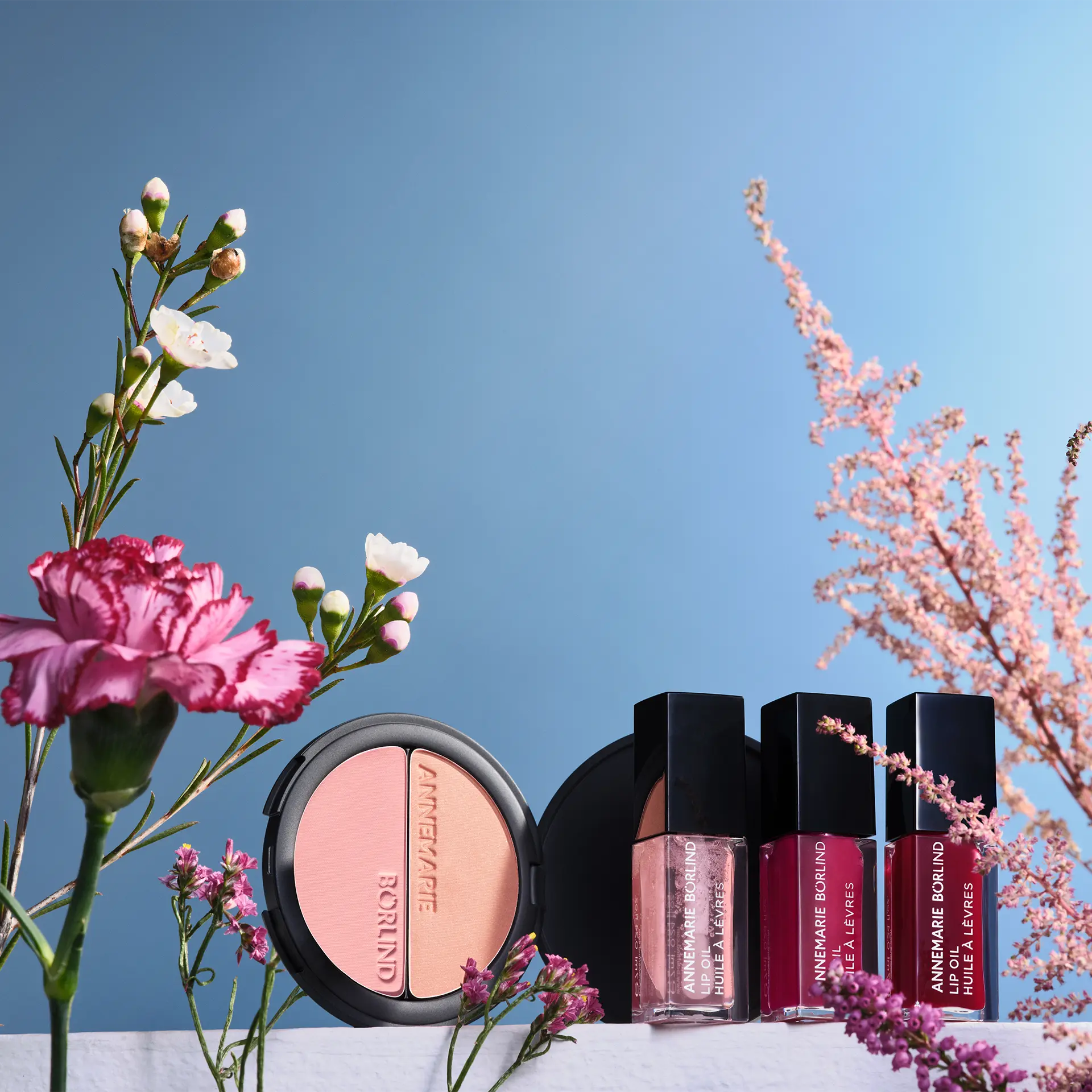 ANNEMARIE BÖRLIND Make-up Duo Blush & Eye shadow, 3 Lip Oils vor blauem Hintergrund zwischen Blumen