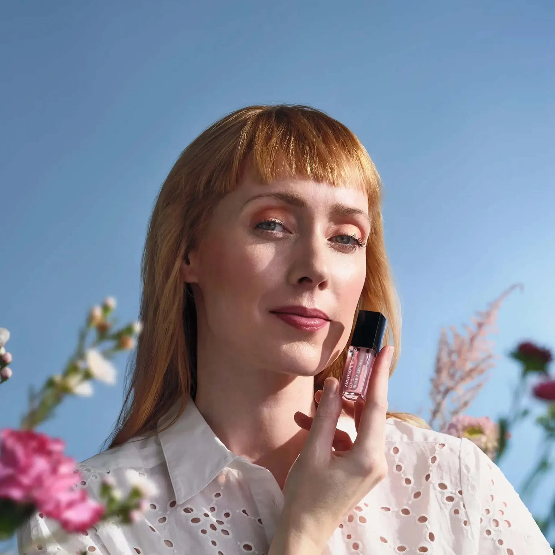 Frau mit frischem Make-up hält das LIP OIL Cherry Blossom von ANNEMARIE BÖRLIND vor blauem Himmel, umgeben von Blüten