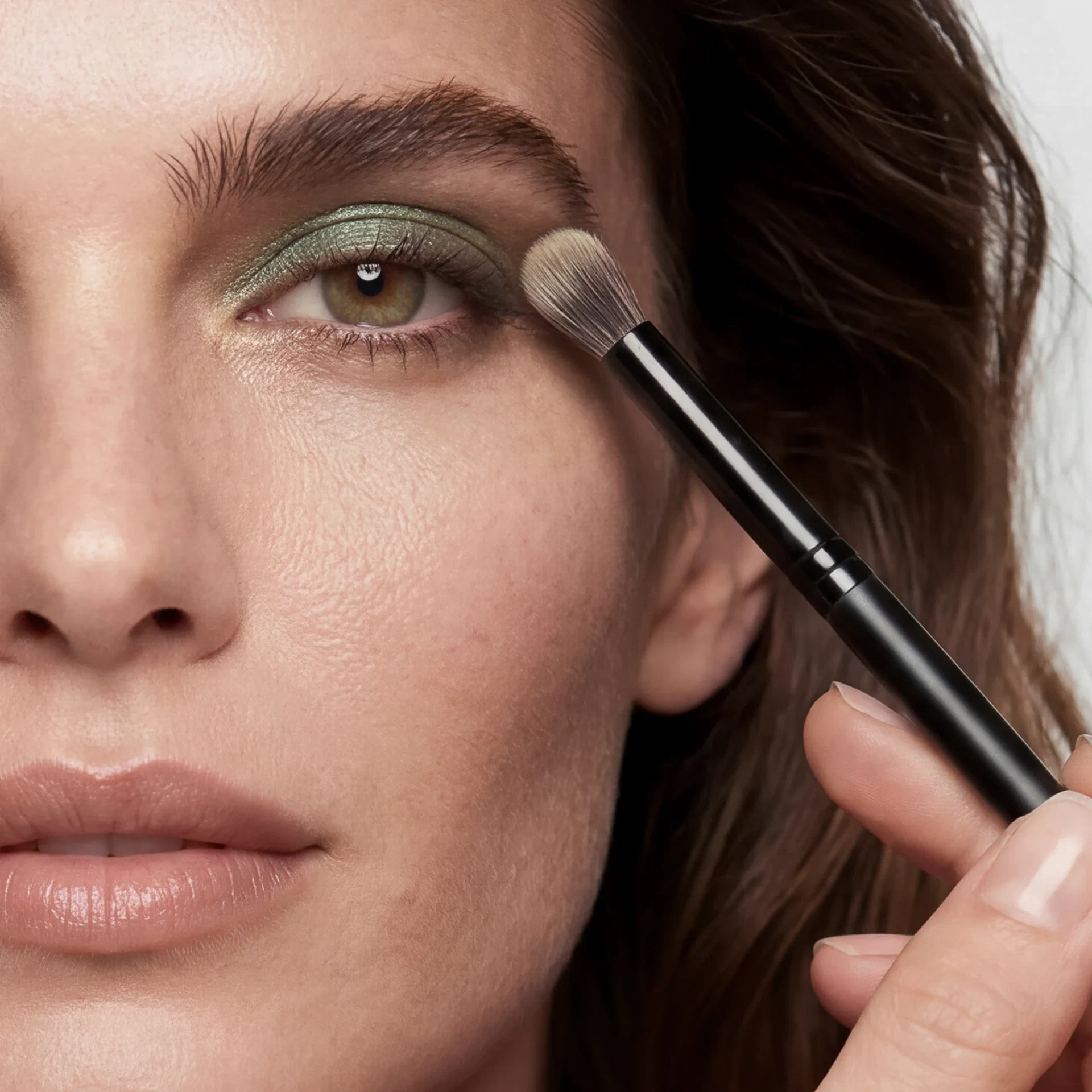 Nahaufnahme eines Auges mit schimmerndem moosgrünem Lidschatten, der mit einem Make-up-Pinsel am äußeren Augenwinkel verblendet wird.