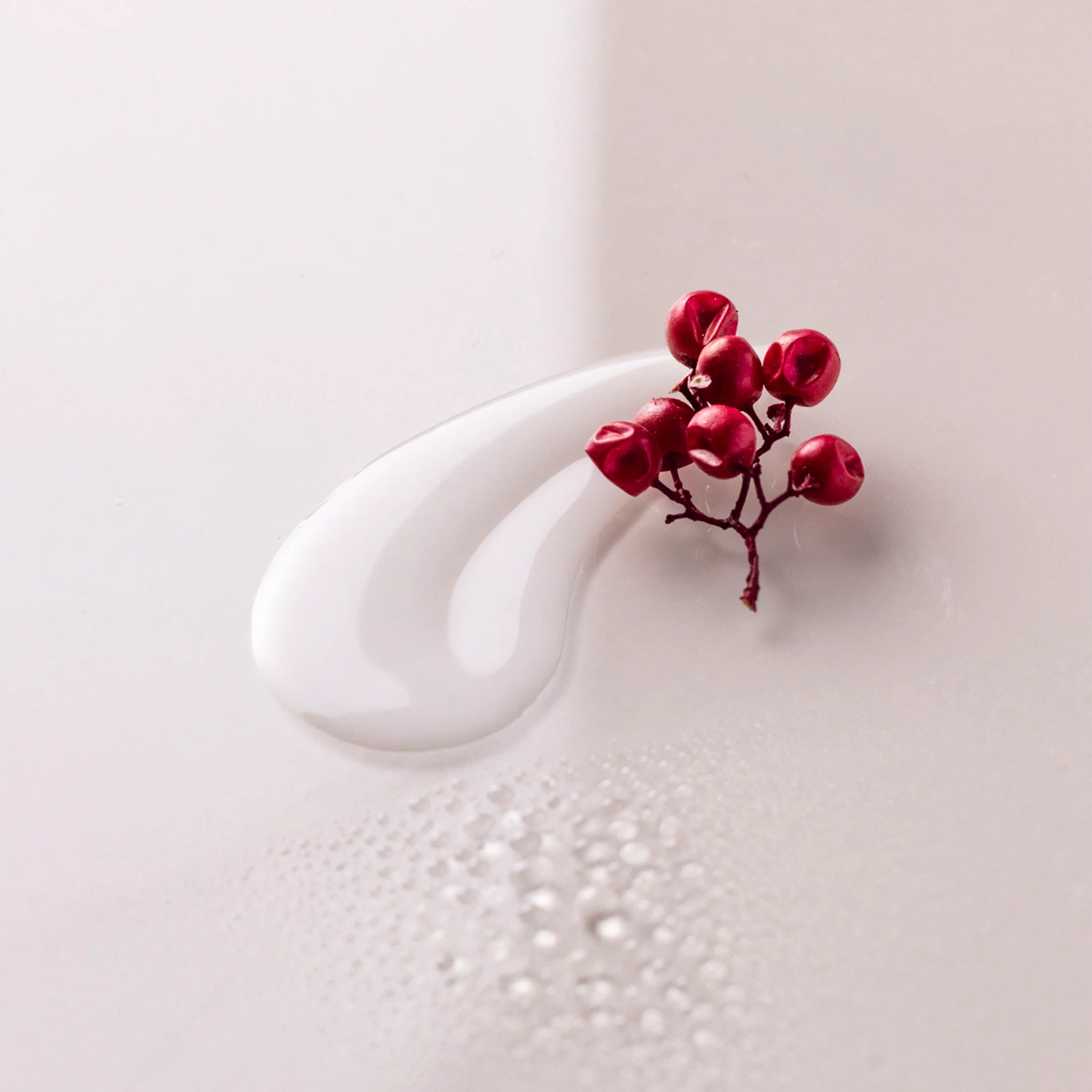Weiße Creme-Textur mit kleinen Wassertropfen und einem Zweig mit roten Beeren auf hellem Untergrund