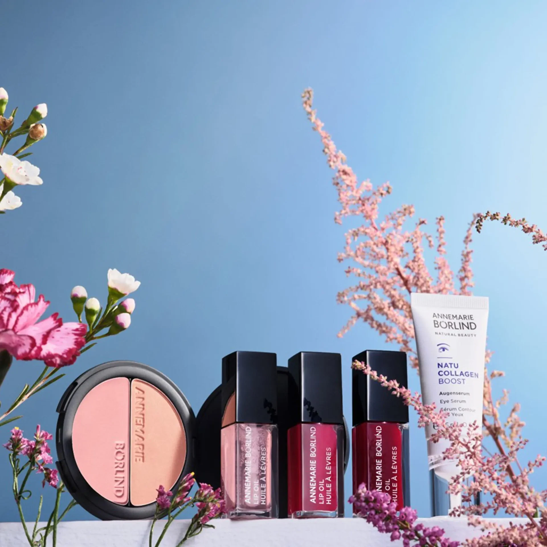 Hier korrigiert:  Mood-Bild 1 (mit Augenserum): SEASON OF BLOOM Kollektion von ANNEMARIE BÖRLIND mit DUO BLUSH & EYE SHADOW, drei LIP OILS und NATUCOLLAGEN BOOST Augenserum, umgeben von Blüten vor blauem Himmel